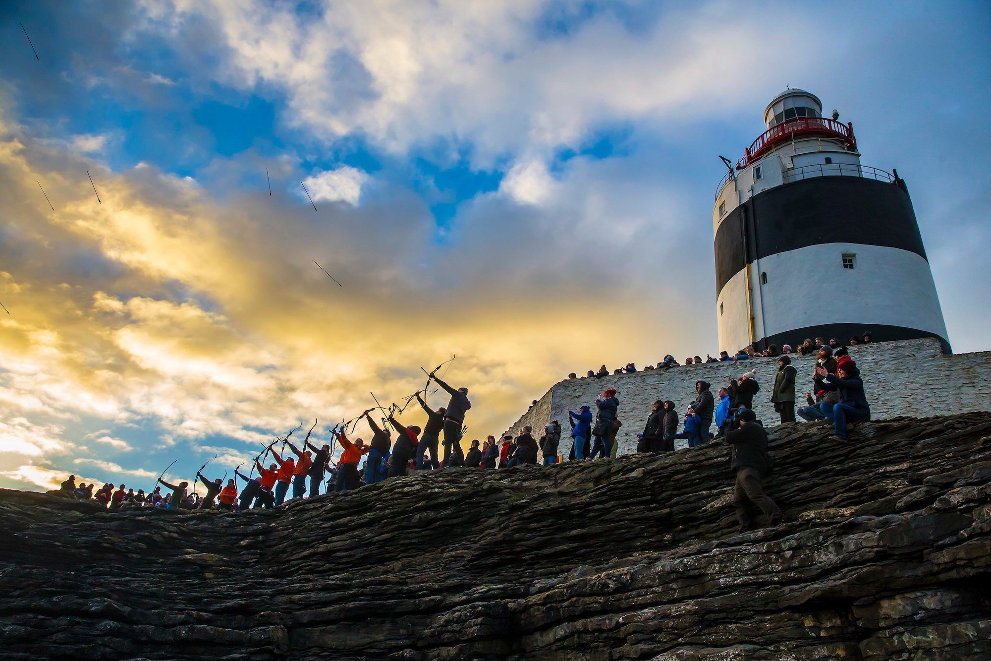 Visitors on the walls beneath the black and white tower of Hook Lighthouse at sunset